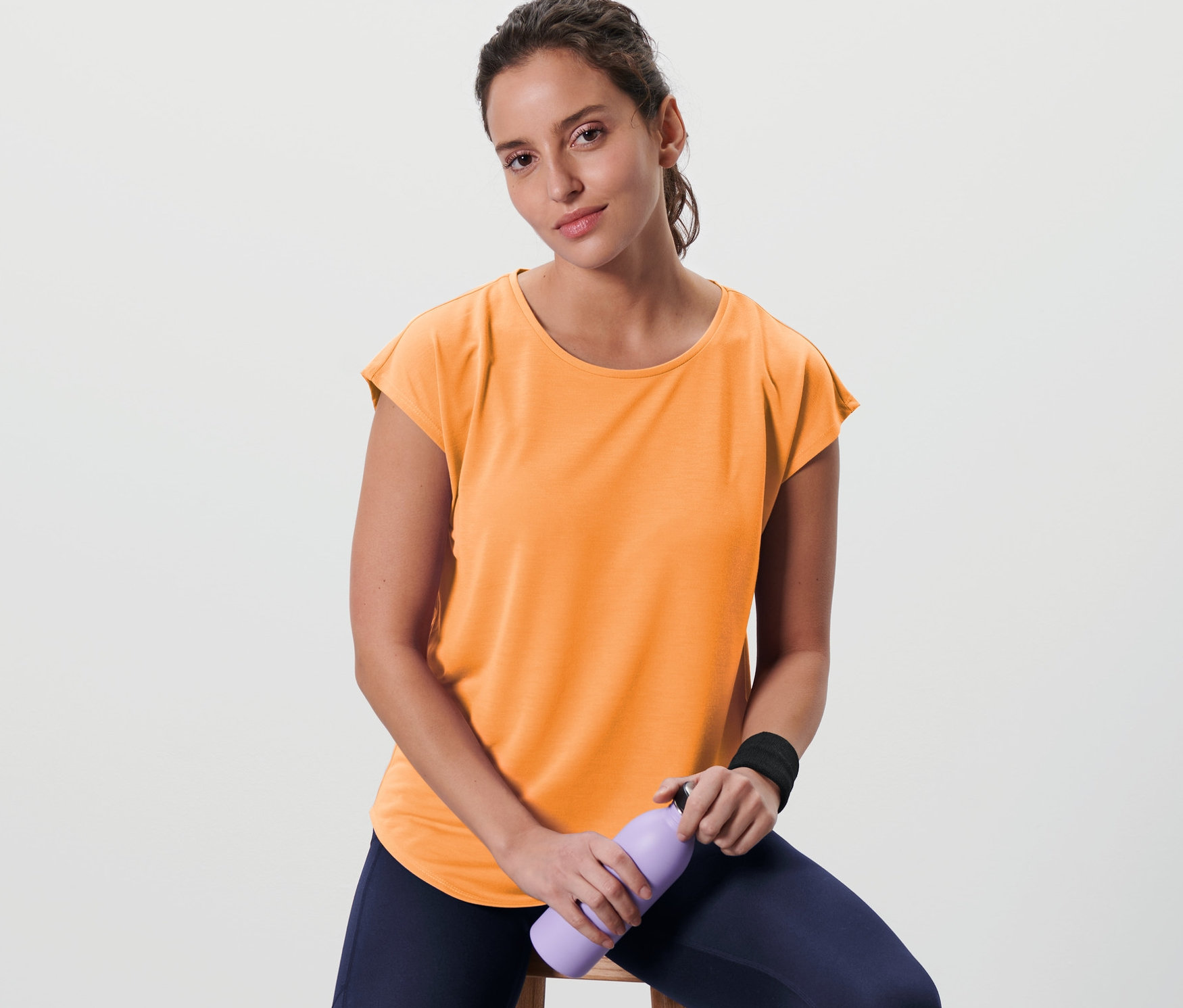 Bir kadın taburede oturuyor ve mor bir su şişesi tutuyor. Şeftali rengi bir spor tişörtü giyiyor.