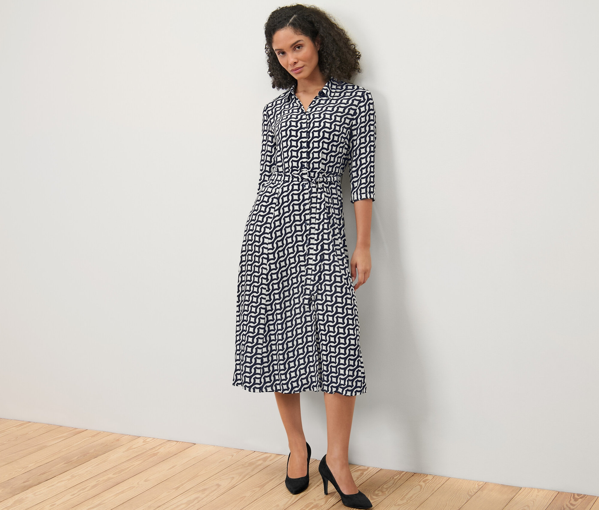 Beyaz bir duvarın önünde duran kadın baskılı bir elbise ve siyah topuklu ayakkabılar giyiyor.