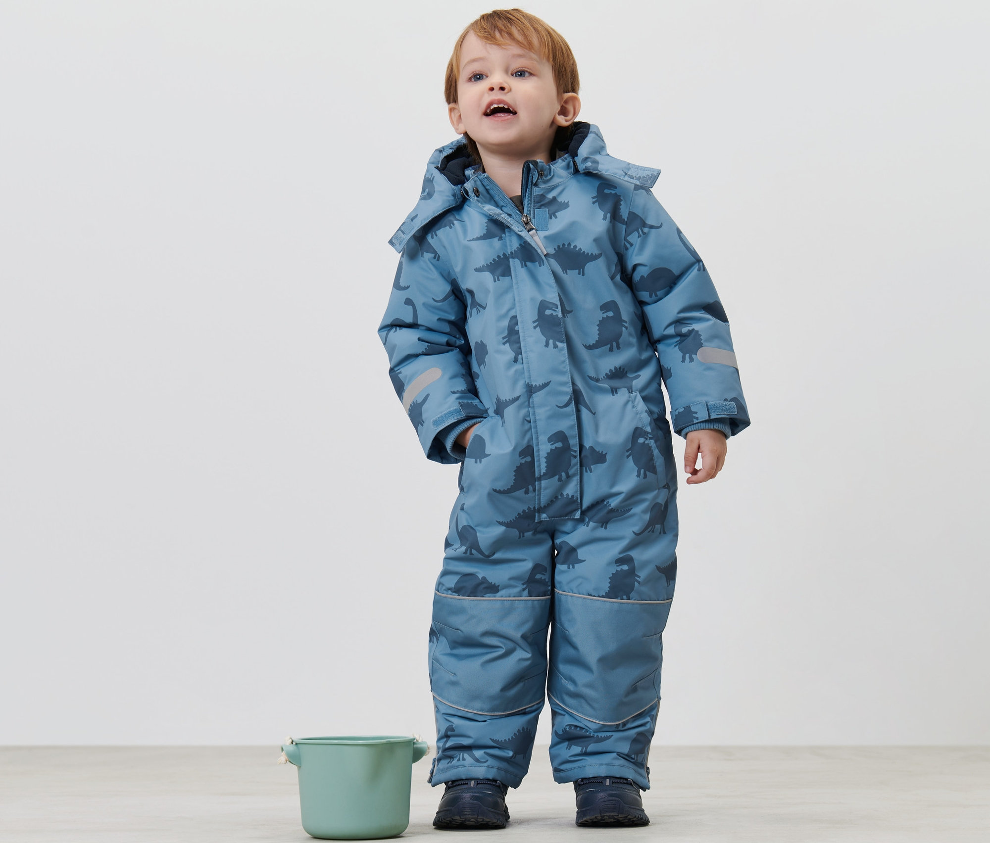 Çocuk, dinozorlu çocuk kar tulumu içinde ayakta duruyor ve yukarı bakıyor. Yanında yeşil bir kova var.