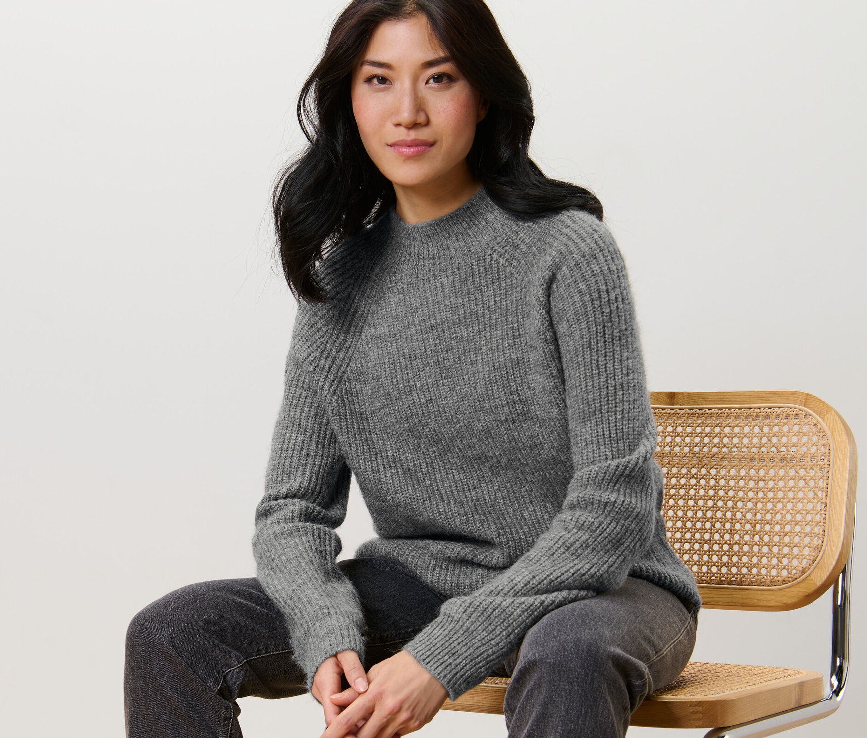 Kadın sandalyede oturuyor ve gri, yarım boğazlı, kalın örgü bir kazak giyiyor.
