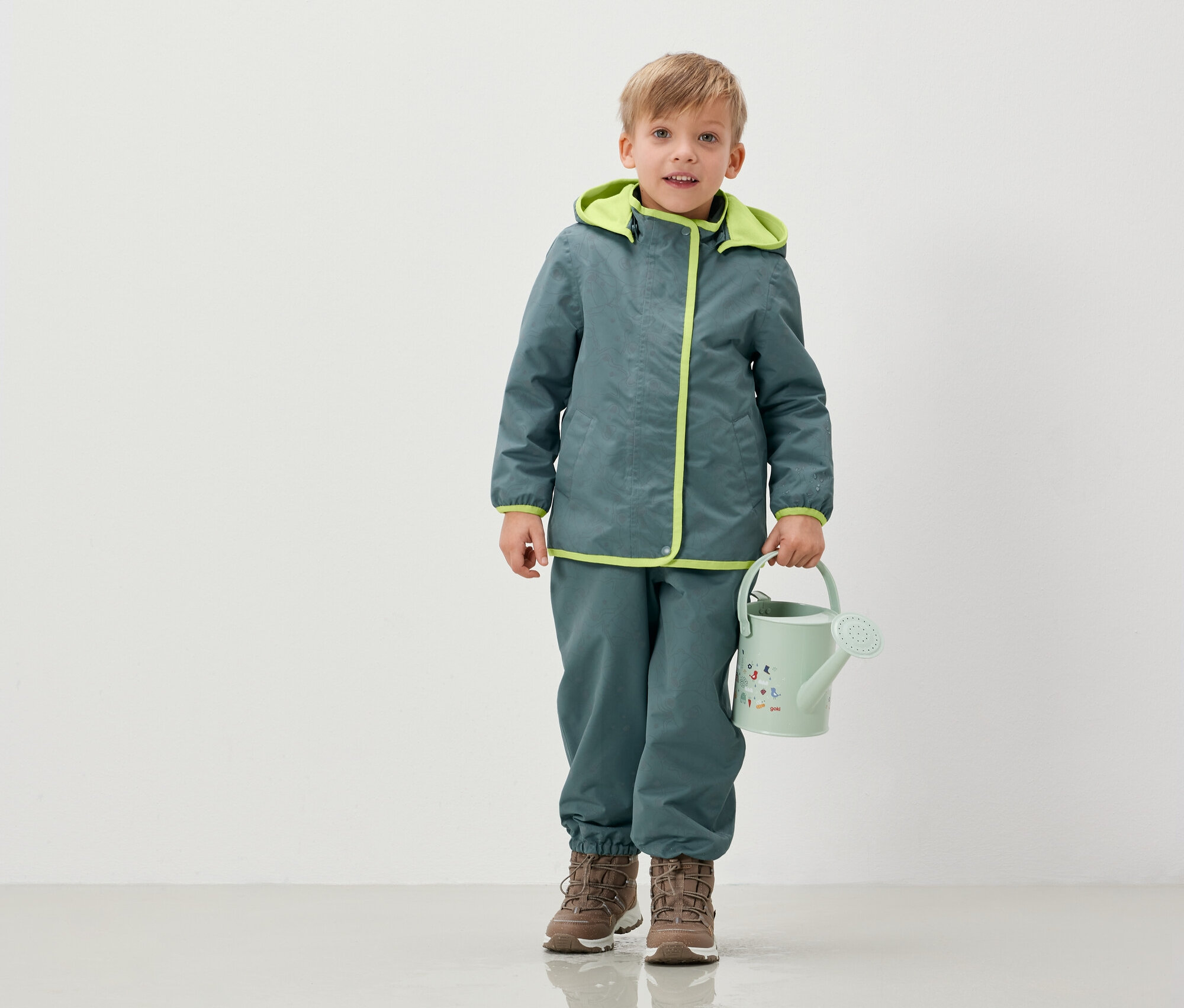 Ceket ve pantolonlu bir çocuk, açık yeşil bir sulama kabı tutuyor.