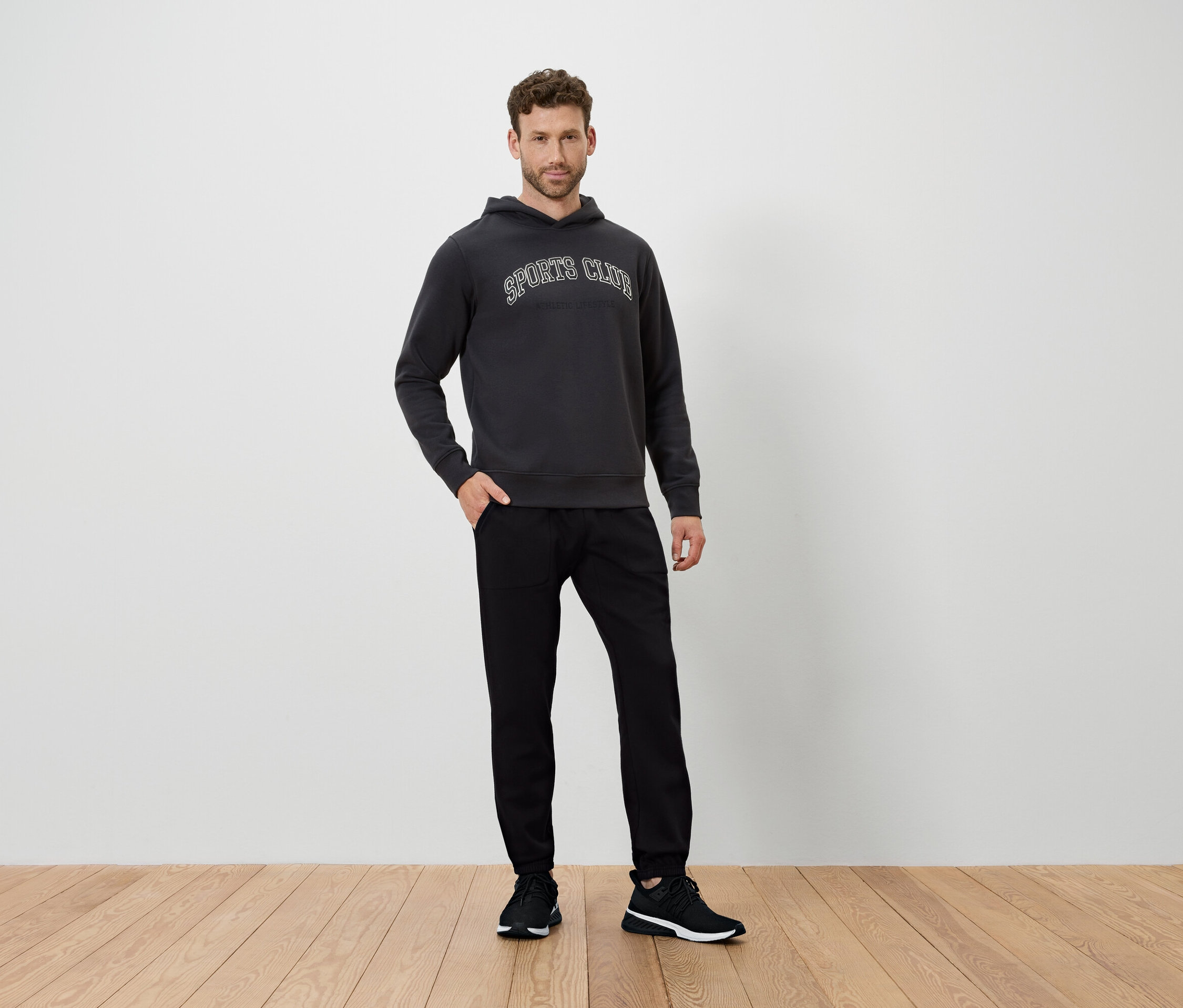 Adam yerde duruyor, koyu gri kapüşonlu ve siyah pantolon giyiyor.