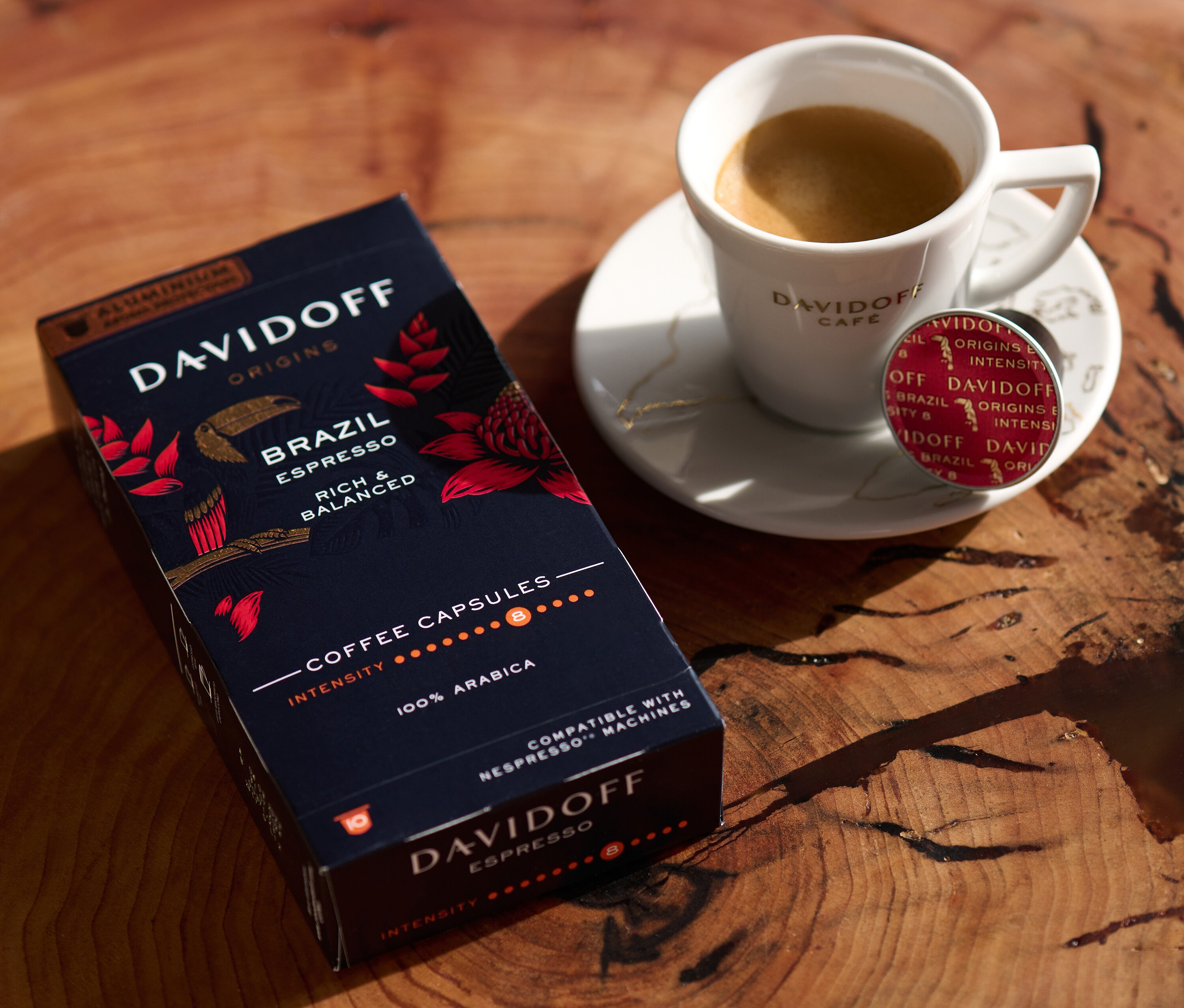 Ahşap bir masanın üzerinde bir paket DAVIDOFF CAFÉ ORIGINS Brazil Espresso, bir fincan espresso ve bir kapsül var.