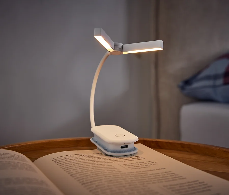 Beyaz, klipsli bir kitap lambası, ahşap bir tepside duran açık bir kitabı aydınlatıyor. Arka planda ekose bir yastık bulunan bir yatak görülüyor.