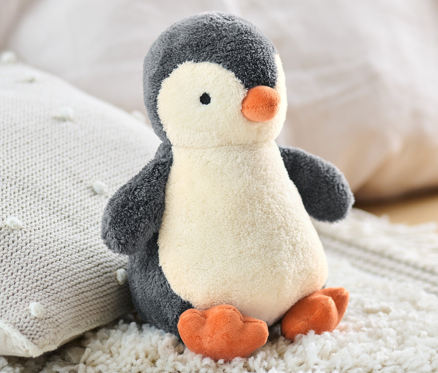 Pelüş Penguen, örgü yastığın yanında beyaz, tüylü bir halının üzerinde oturuyor.