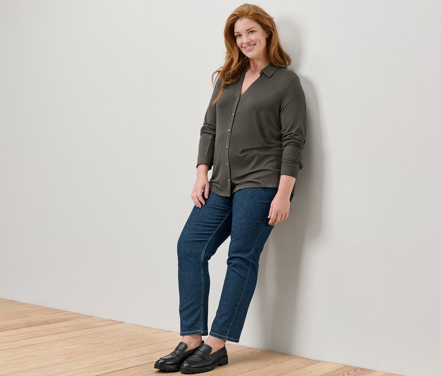 Duvara yaslanmış bir kadın, Düğmeli Bluz ve kot pantolon giyiyor.