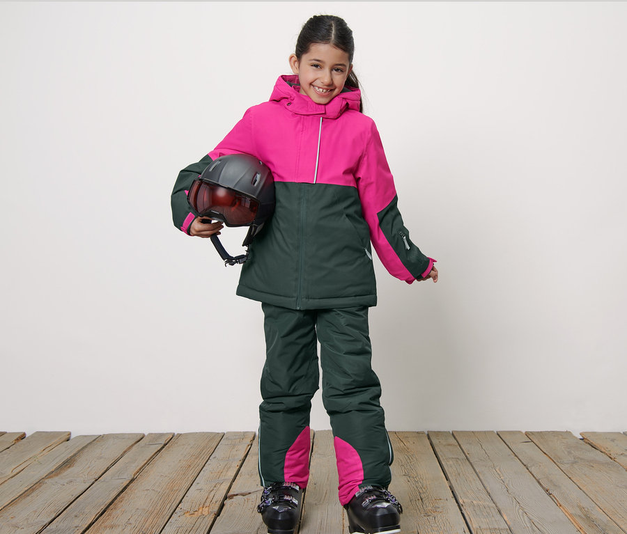 Çocuk kayak montu giyen gülümseyen bir kız çocuğu kask tutuyor.