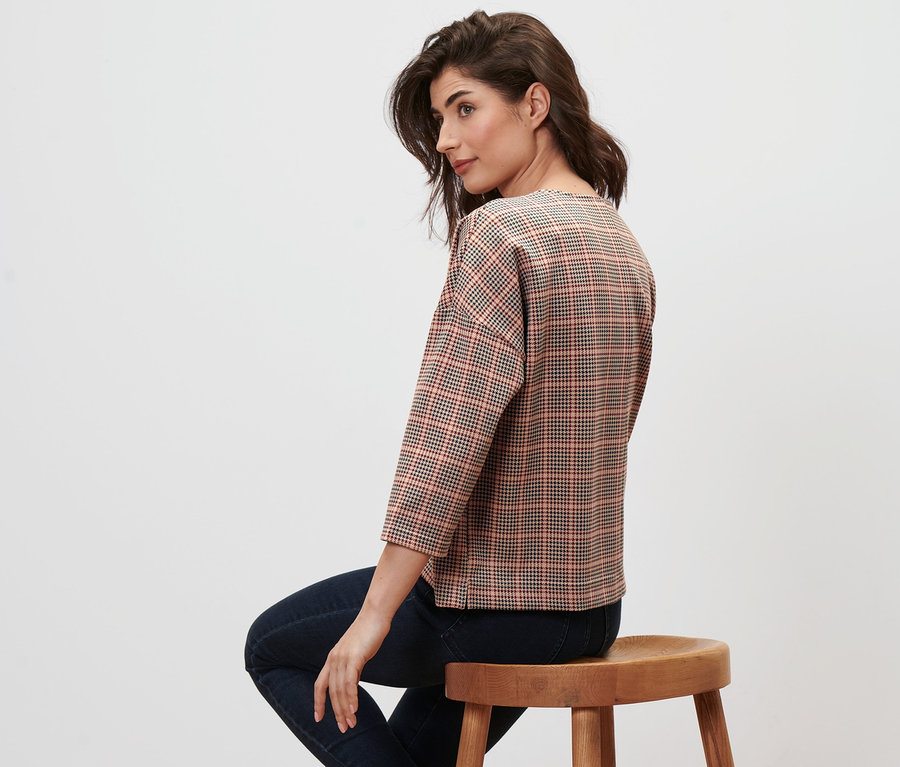 Kadın ahşap bir taburede oturuyor ve 3/4 kollu, kazayağı desenli bir sweatshirt giyiyor.