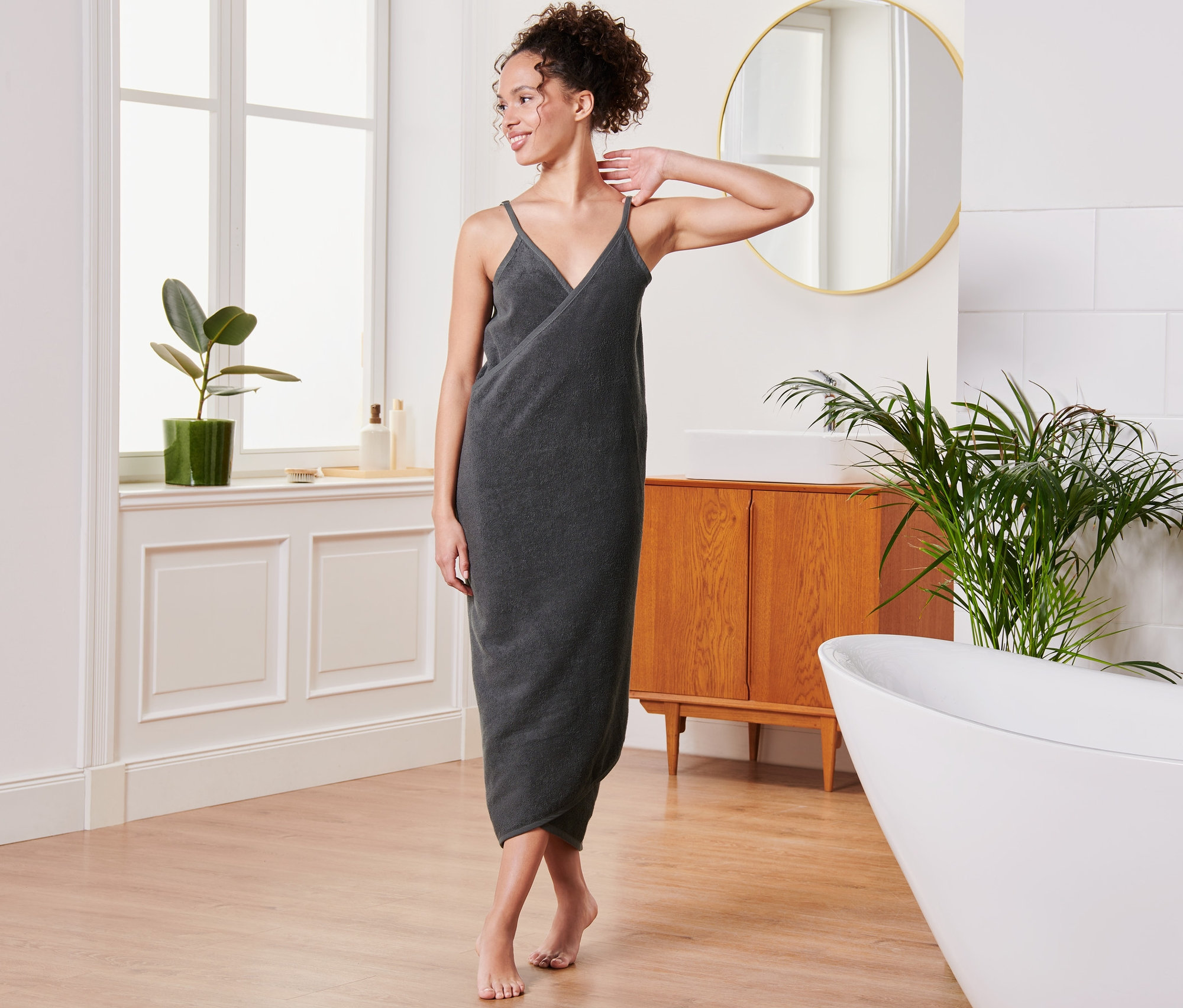 Bir kadın banyoda antrasit havlu elbise içinde poz veriyor.