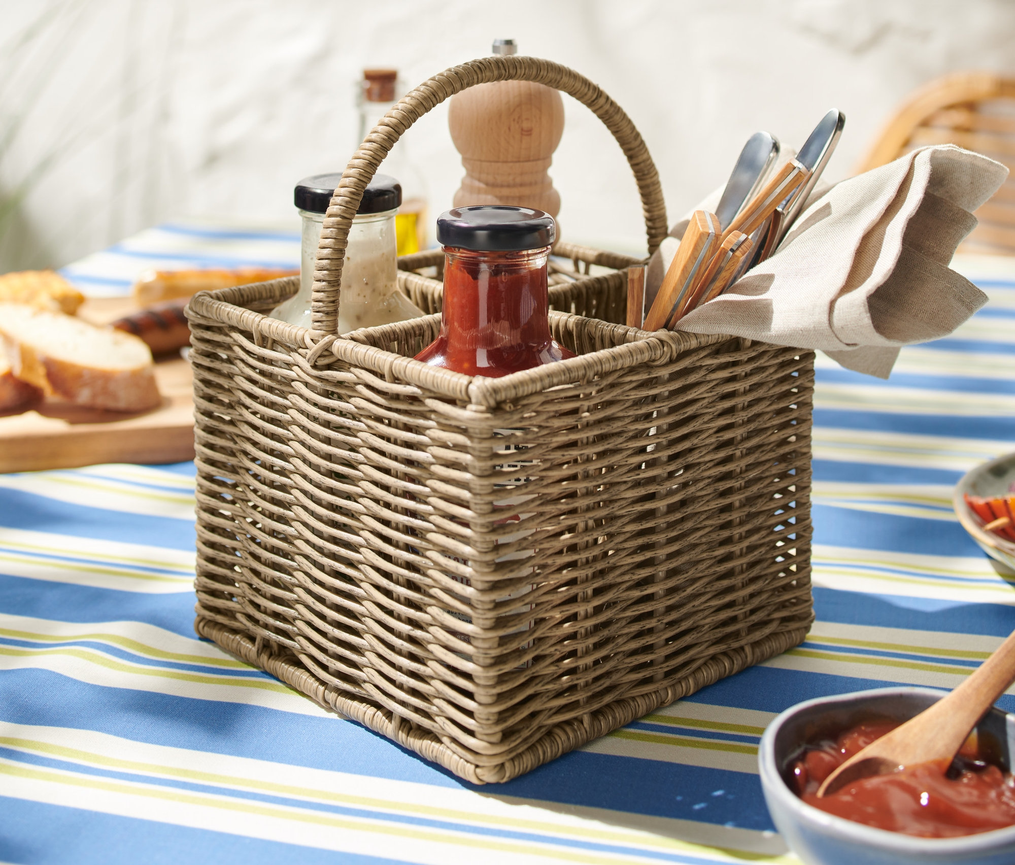 Mavi beyaz çizgili masa örtüsünün üzerinde duran hasır servis sepeti. Sepetin içinde bardaklar, karabiber değirmeni, peçete ve servis takımları bulunur.