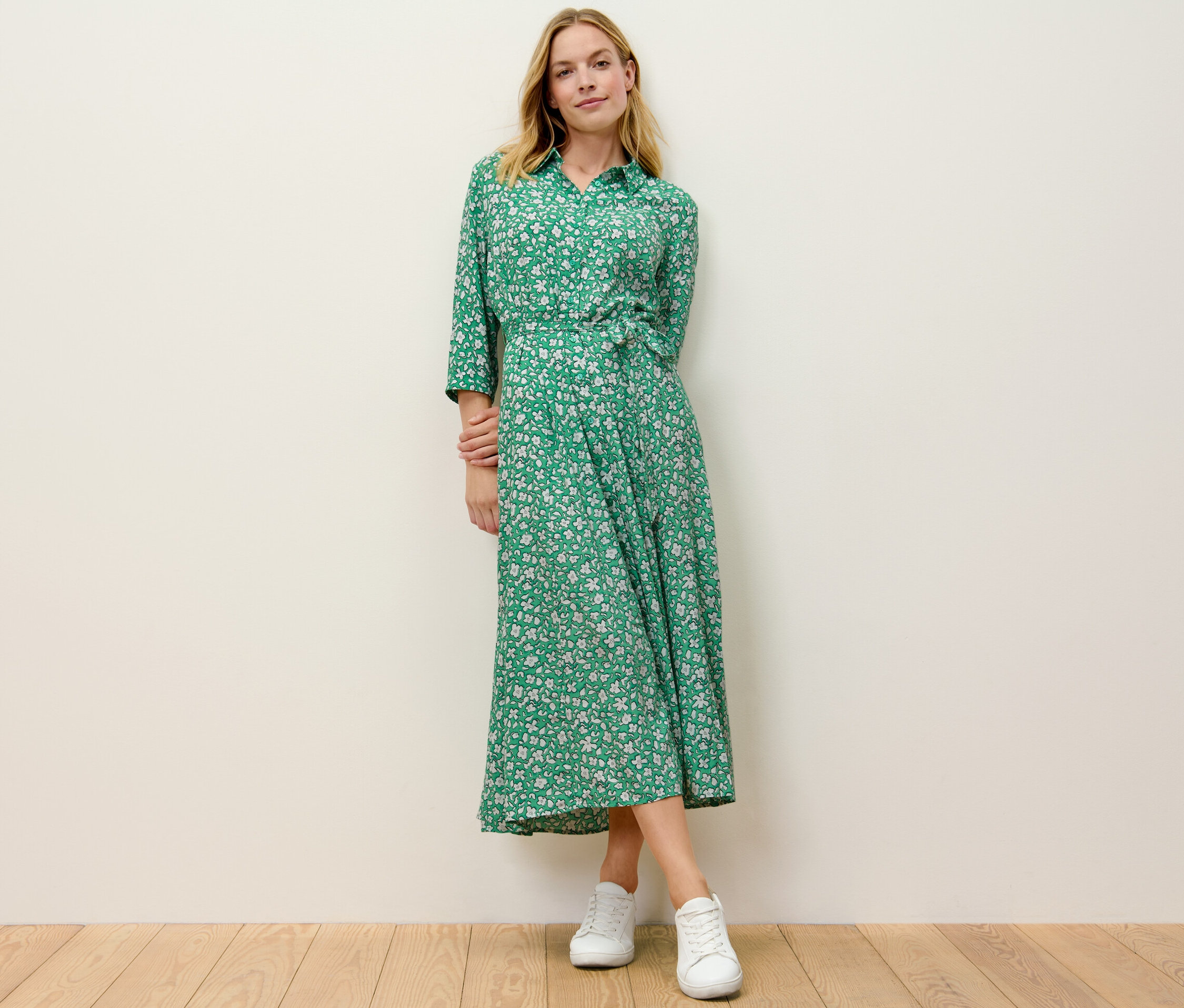 Sarışın bir kadın ayakta duruyor ve baskılı, yeşil bir elbise giyiyor.