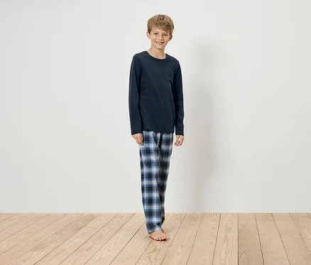 Ahşap zeminde duran, lacivert uzun kollu bir pijama üstü ve mavi kareli bir pijama altı giyen bir erkek çocuk. 