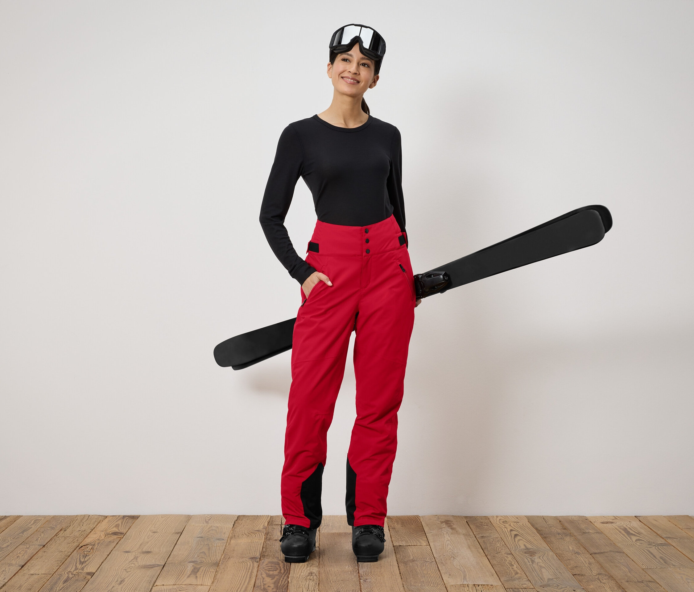 Kadın ayakta duruyor ve kayak tutuyor. Siyah uzun kollu bir gömlek, kırmızı pantolon ve kayak gözlüğü takıyor.