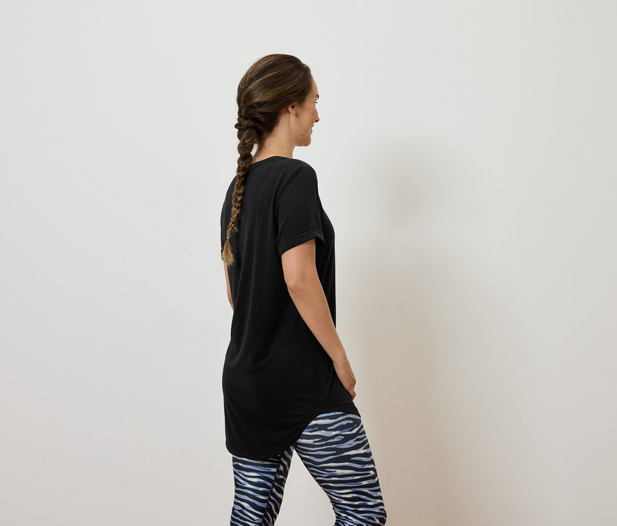 Kahverengi saçları örgülü bir kadın, sırtı kameraya dönük bir şekilde duruyor, üzerinde siyah bir tişört ve mavi beyaz tayt var.