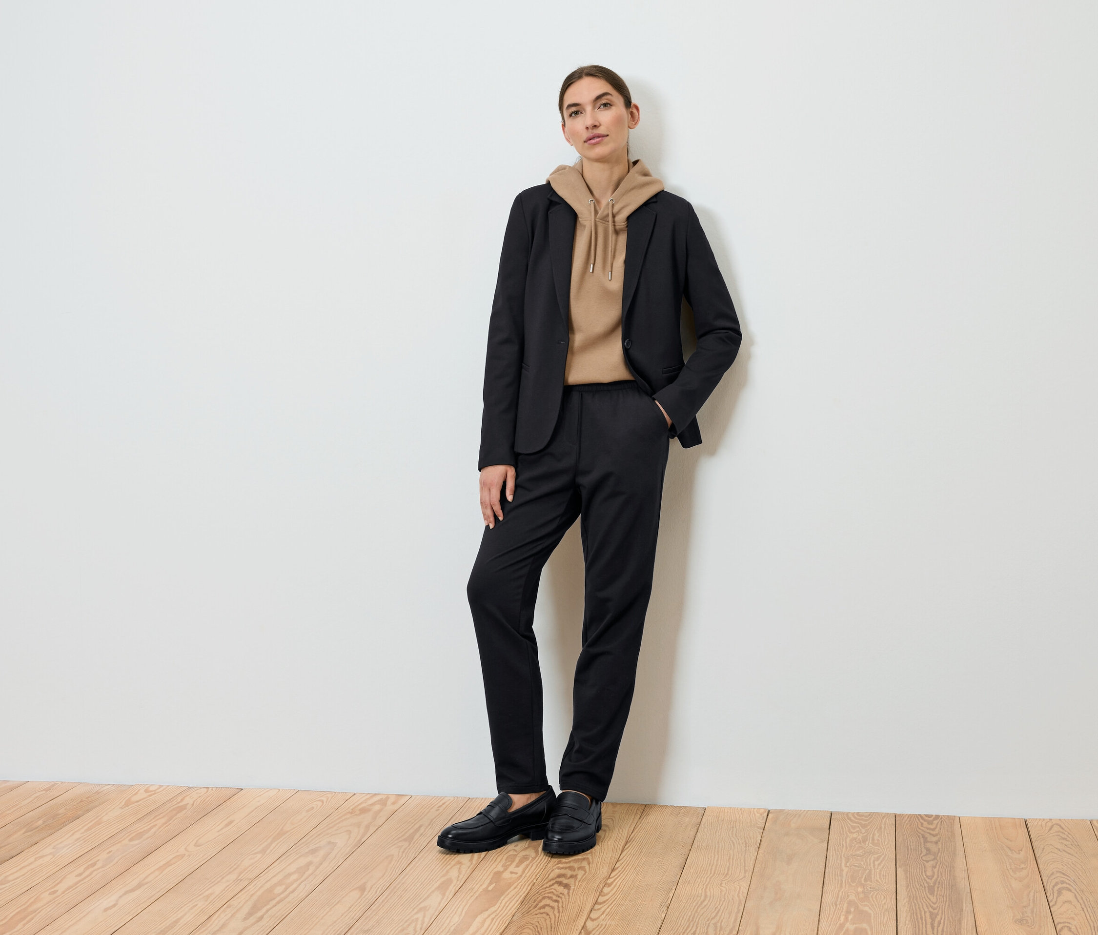 Bir kadın siyah bir blazer, kahverengi bir kapüşonlu sweatshirt ve siyah pantolon giyerek poz veriyor.