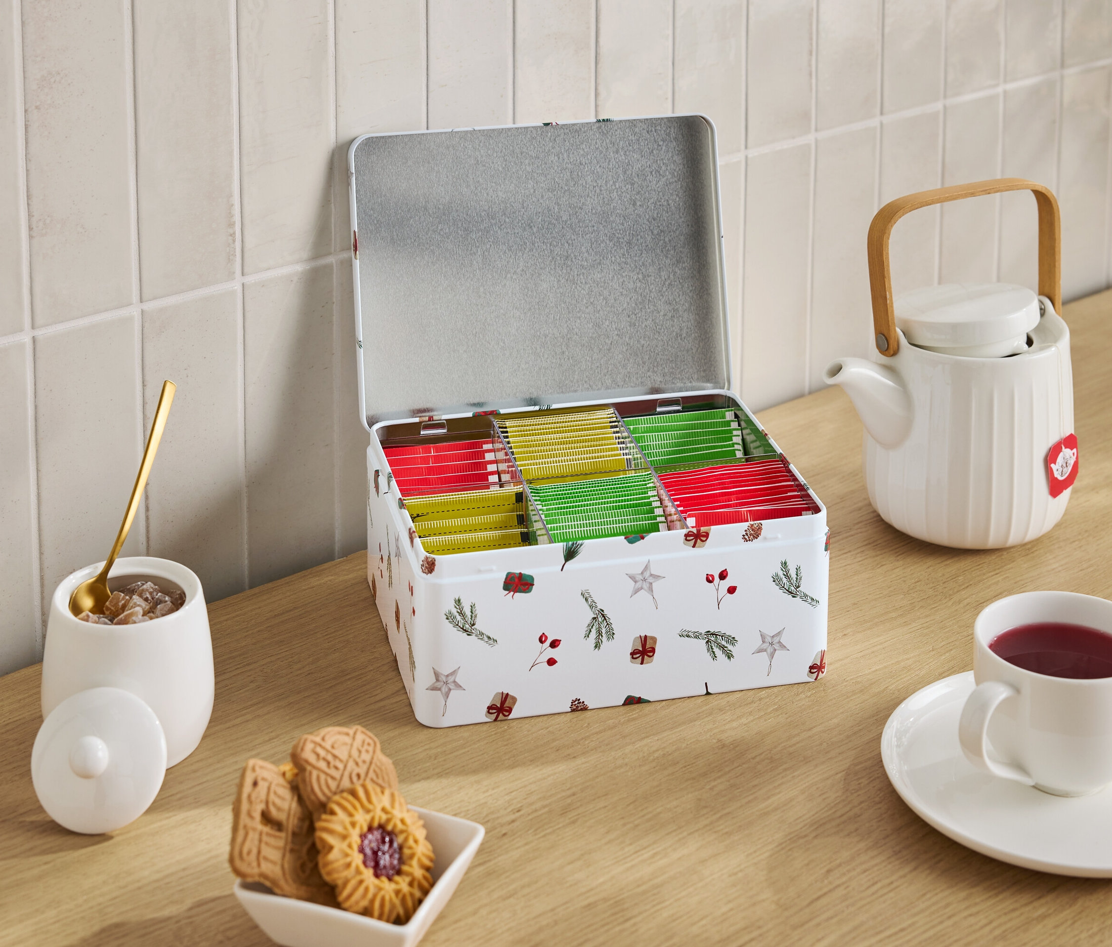 Noel temalı, açık bir çay kutusu çay poşetleriyle dolu, beyaz bir çaydanlık, bir fincan çay, şekerli bir şekerlik ve bir kase kurabiyenin yanında duruyor.