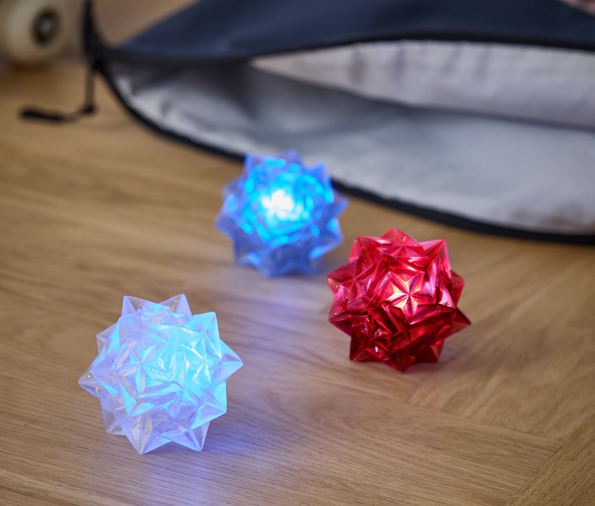 Ahşap bir zeminde üç adet ışıklı top duruyor: beyaz, mavi ve kırmızı. Arkalarında bir sırt çantası ve kaykay var.