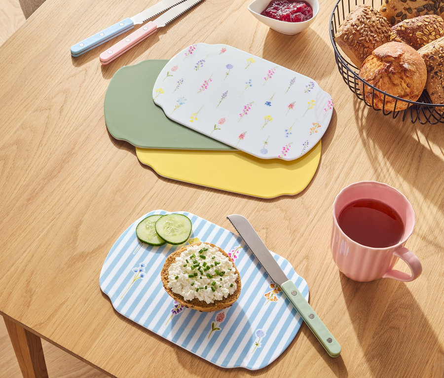 Ahşap bir masa üzerinde bir sepette ekmekler, reçel, bıçaklar ve farklı renklerde kesme tahtaları bulunmaktadır.