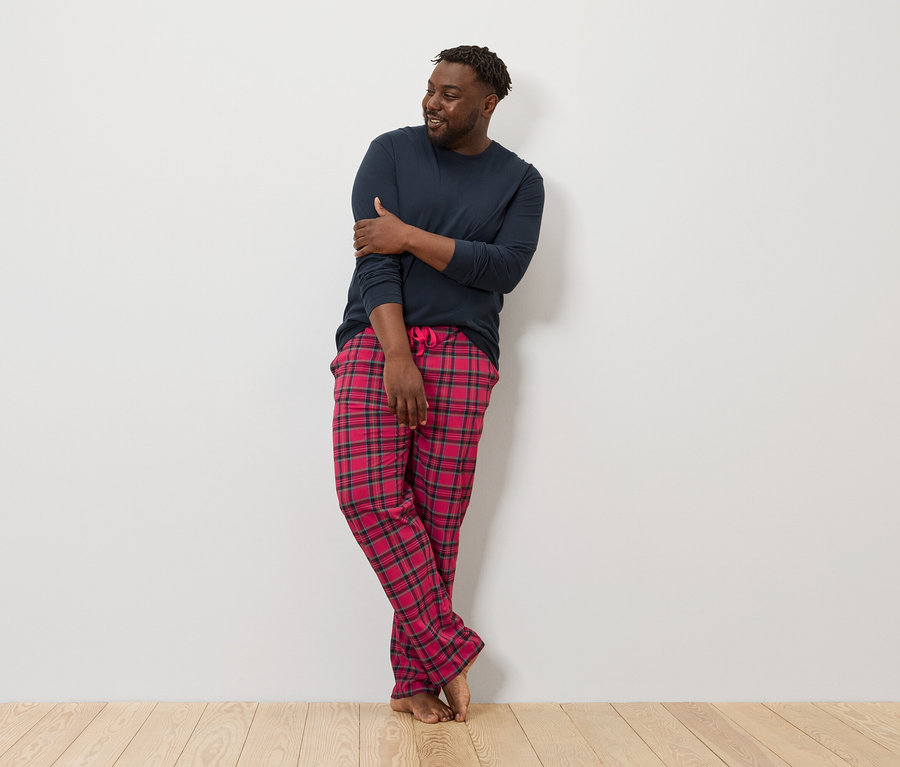 Duvara yaslanmış bir adam lacivert uzun kollu bir tişört ve kırmızı ekose bir pijama altı giyiyor.
