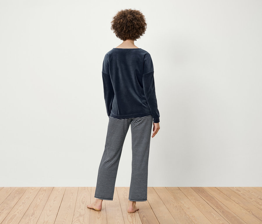 Kıvırcık saçlı bir kadın arkası dönük duruyor, lacivert kadife bir üst ve çizgili bir pantolon giyiyor.