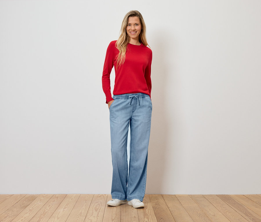 Kırmızı bir kazak ve açık mavi, İspanyol paça bir pantolon giyen bir kadın ahşap bir zeminde duruyor.