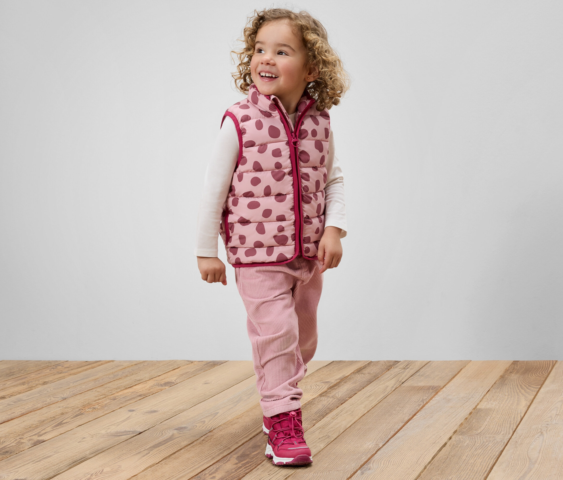 Kıvırcık saçlı, pembe benekli yelek, pembe pantolon ve pembe ayakkabılar giyen gülümseyen bir kız çocuğu ahşap bir zeminde duruyor.