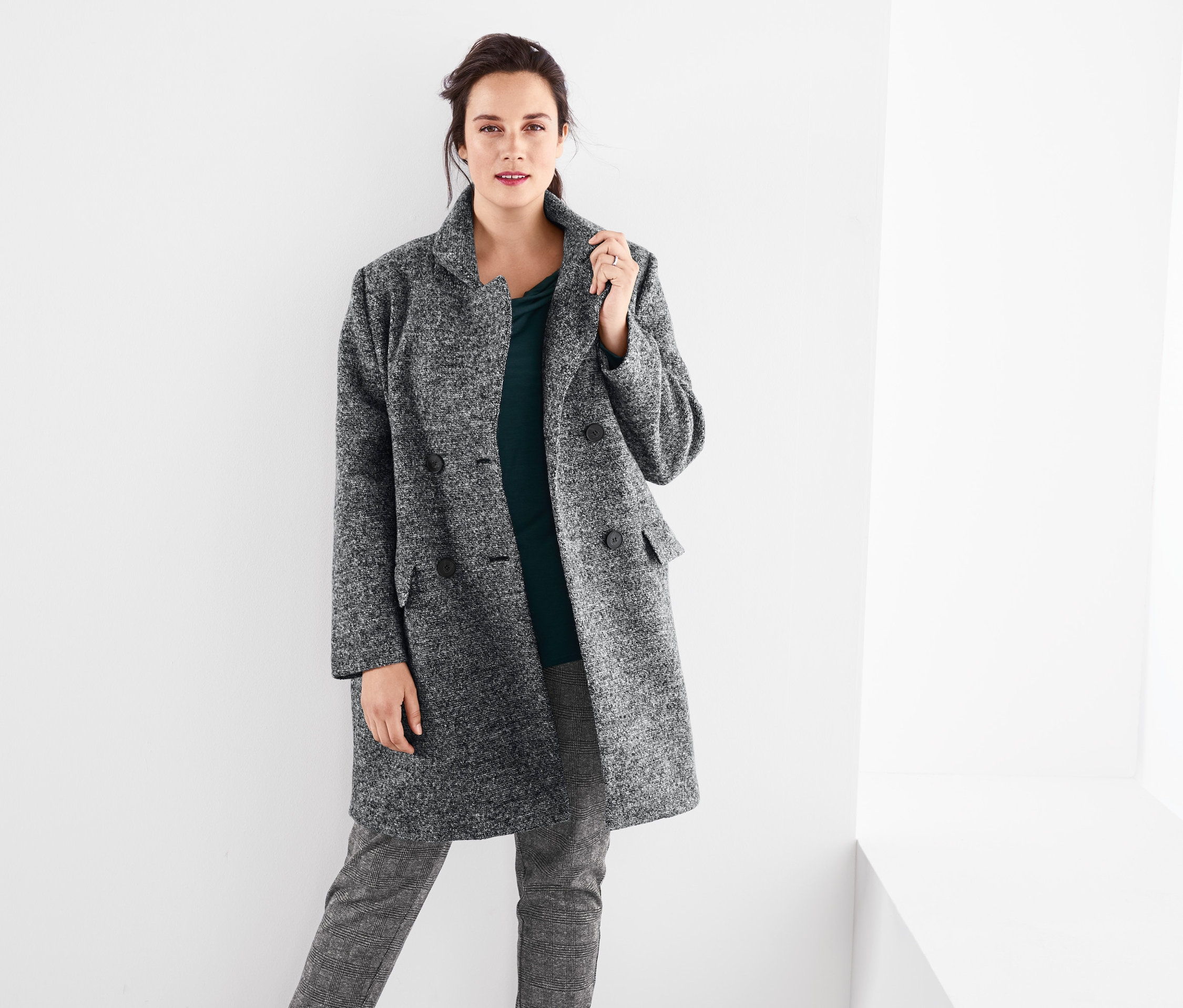 Kadın kırçıllı gri palto, dökümlü tişört ve kareli rahat pantolon giyiyor.