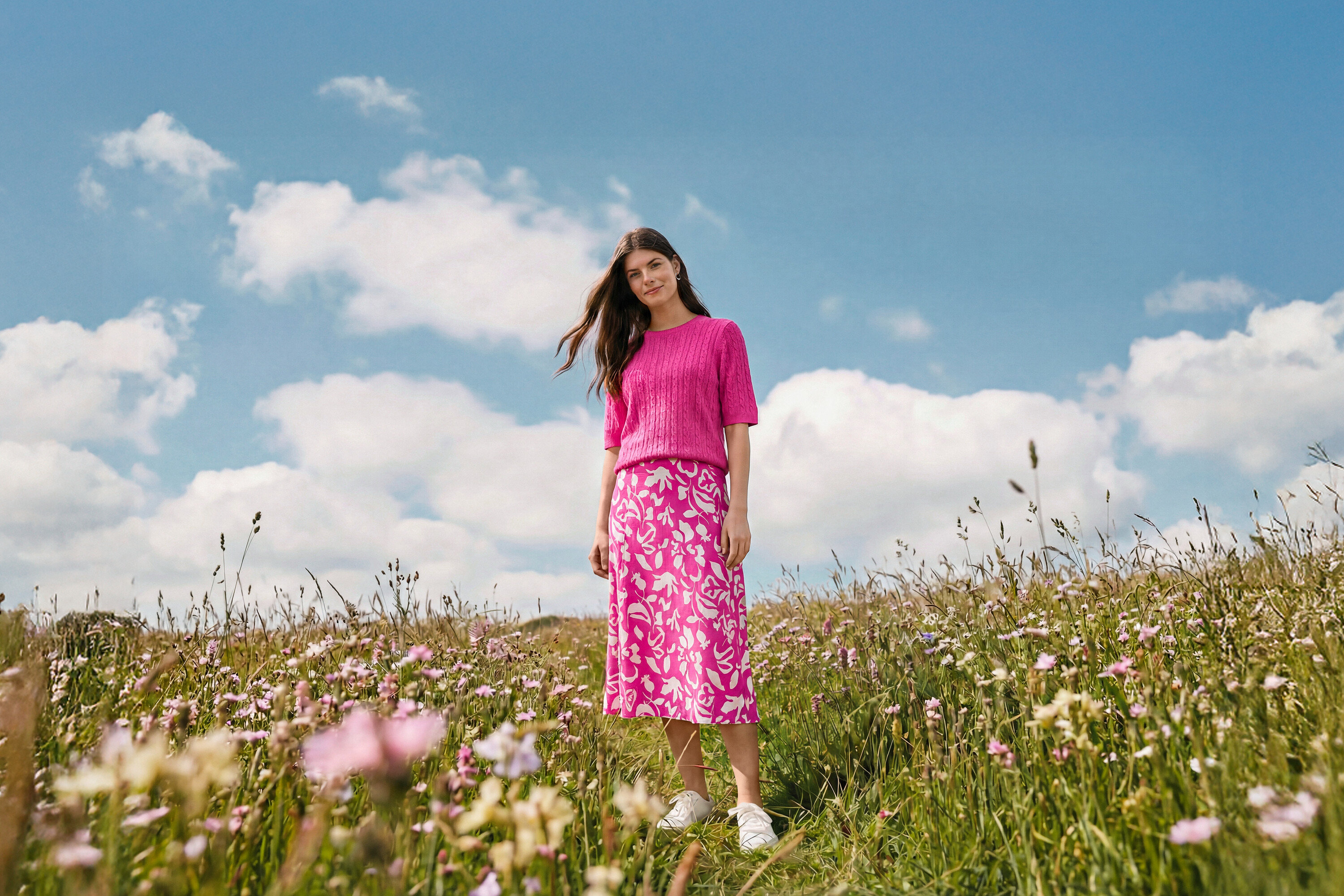 Çayırda duran bir kadın, üzerinde baskılı midi etek, Dalga Halka Küpe ve deri spor ayakkabı var.