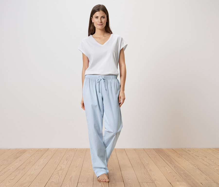 Bir kadın ahşap bir zeminde çıplak ayakla duruyor, beyaz bir tişört ve açık mavi pijama altı giyiyor.