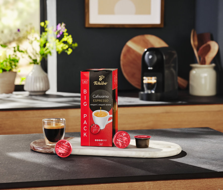 Masanın üzerinde bir paket Cafissimo Espresso kapsülü, bir bardak espresso ve bir kahve makinesi duruyor.
