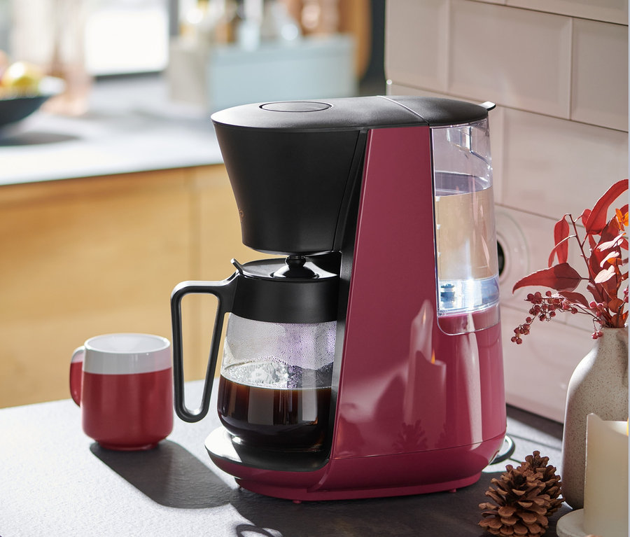 Berry renkli »Let's Brew« filtre kahve makinesi, mutfak tezgahında kırmızı bir kahve fincanının yanında duruyor.