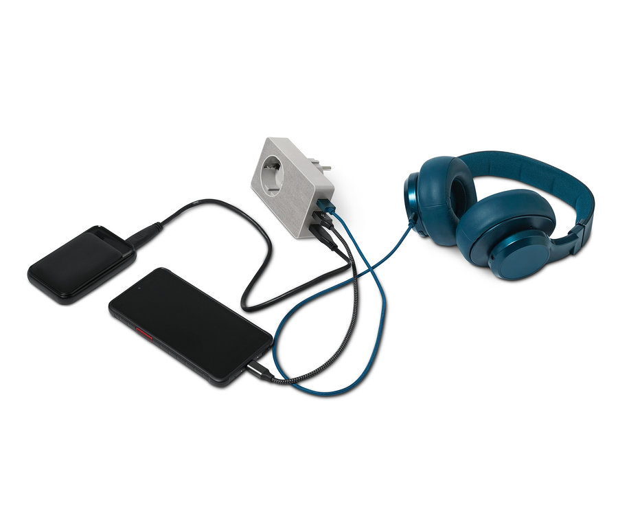 Siyah bir akıllı telefon ve powerbank, 4'ü 1 arada USB priz adaptörü ile şarj ediliyor. Adaptörün yanında mavi kulaklıklar bulunuyor.