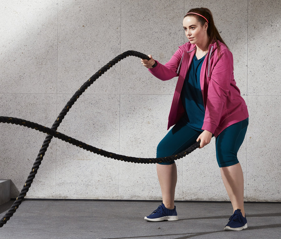 Kadın kırçıllı pembe kapüşonlu sweat hırka ve mavi spor tişörtü ile halatlarla egzersiz yapıyor.