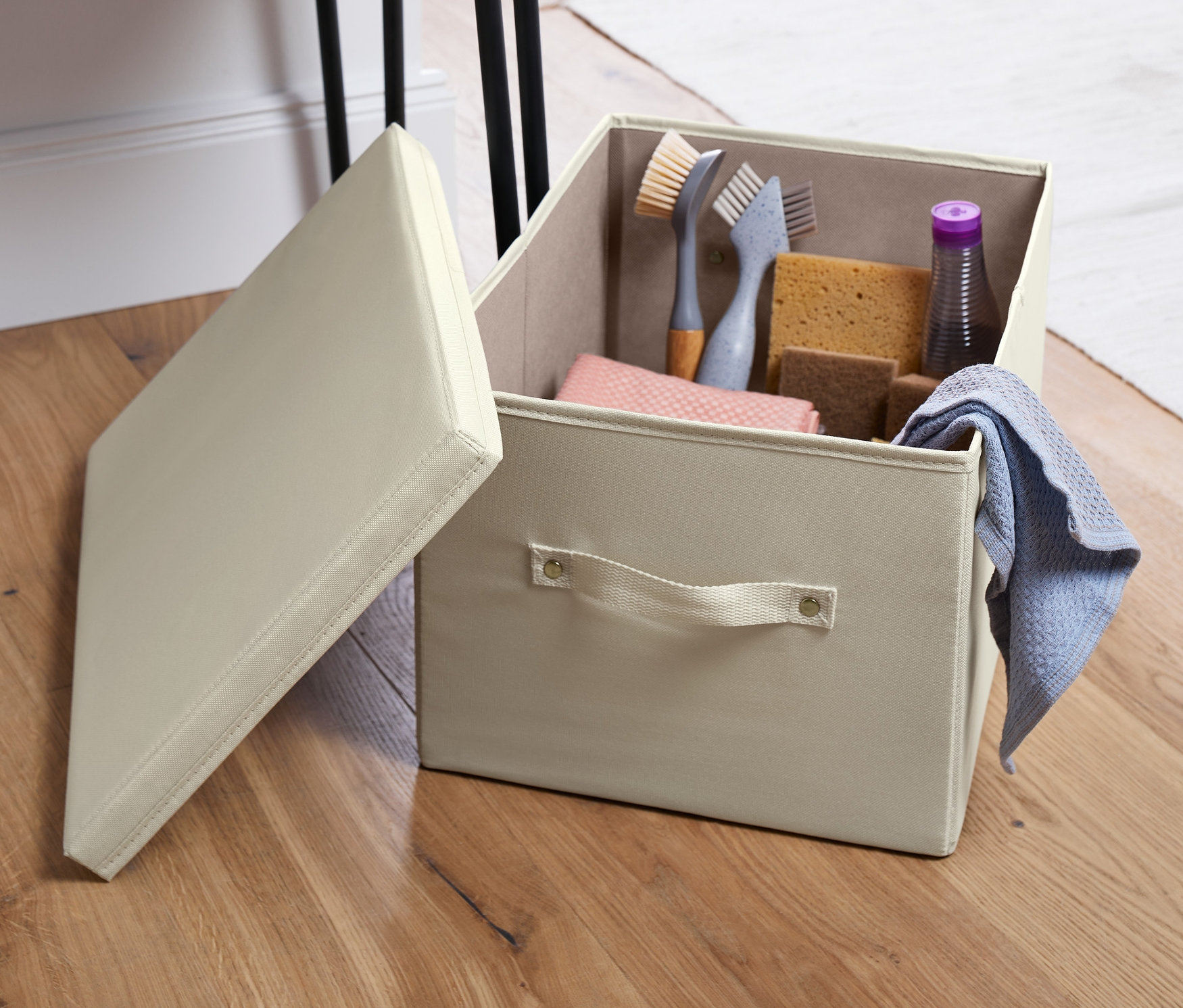 Açık, krem rengi kapaklı düzenleme sandığı, temizlik ürünleriyle dolu, ahşap bir zeminde masanın yanında duruyor.