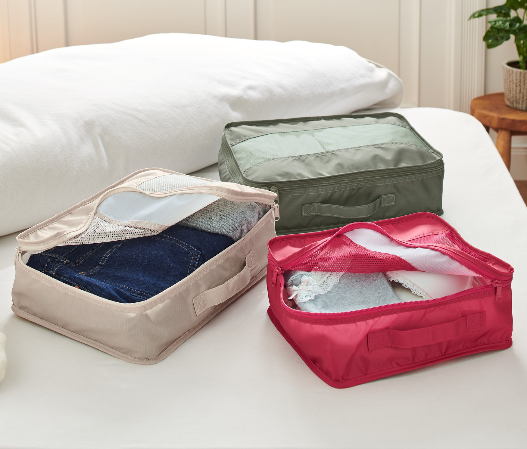 Bir yatak üzerinde her biri farklı renk ve içeriğe sahip üç adet Seyahat Düzenleme Çantası bulunmaktadır.