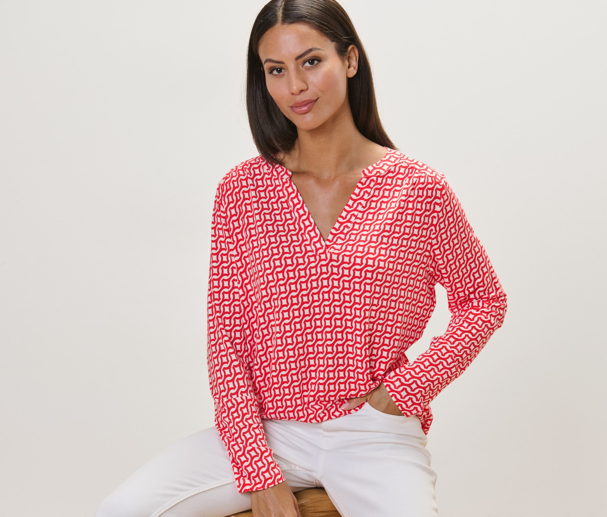 Kadın taburede oturuyor, üzerinde kırmızı baskılı bluz var.