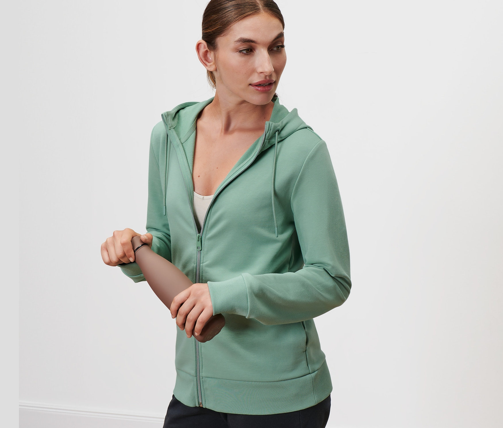 Kadın kahverengi bir su şişesi tutuyor ve Ada Çayı Yeşili renginde bir Sweat Ceket giyiyor.