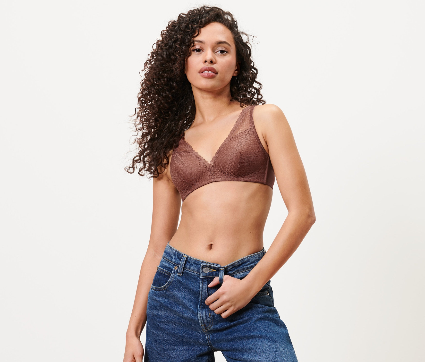 Kadın kestane rengi yumuşak kuplu sütyen ve mavi kot pantolonla poz veriyor.