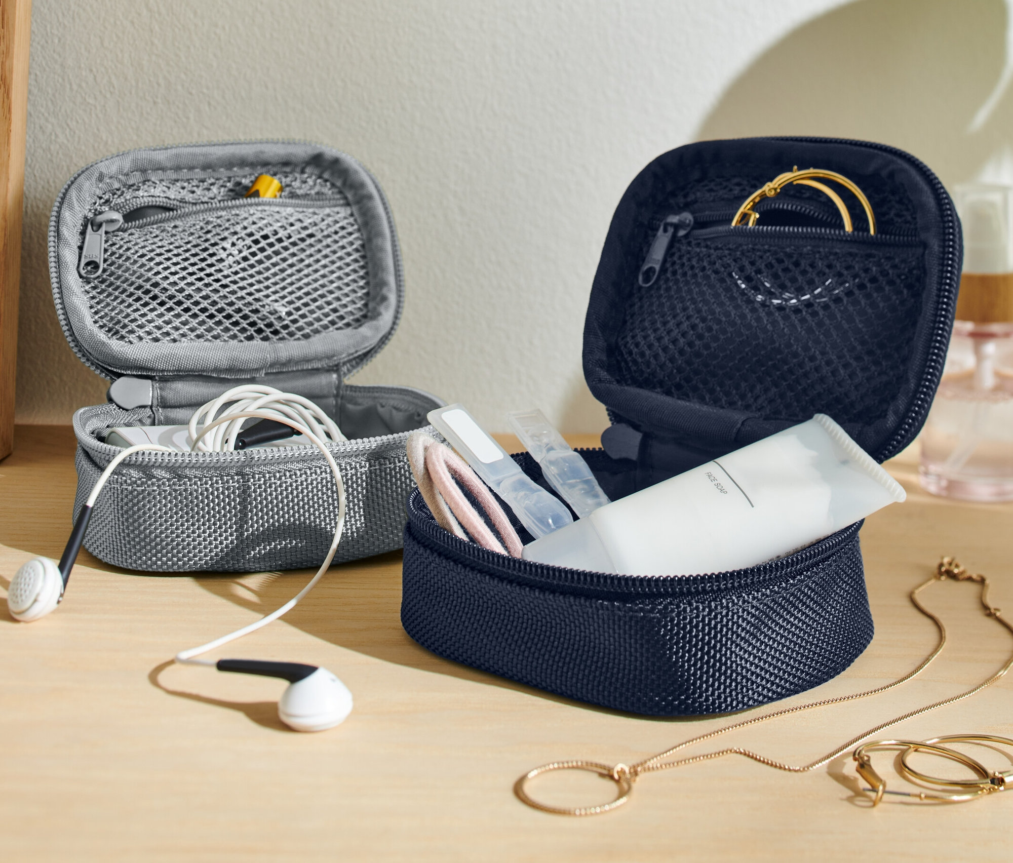 İki adet açık mini seyahat düzenleme çantası; biri beyaz kulaklıklı gri, diğeri kozmetik ve altın takılarla dolu lacivert renkte.