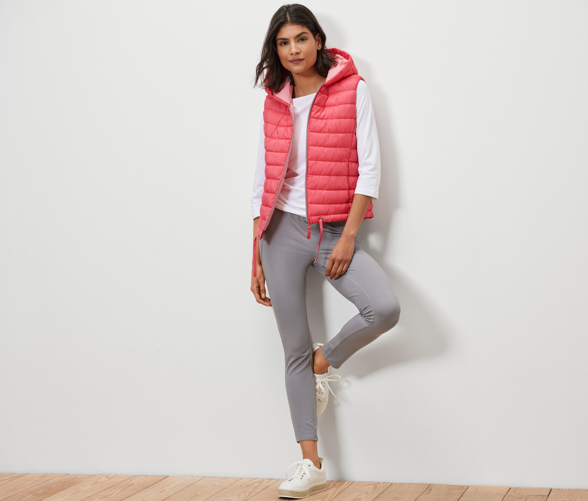 Kadın kapüşonlu kapitone yelek, beyaz tişört ve gri pantolonla poz veriyor.