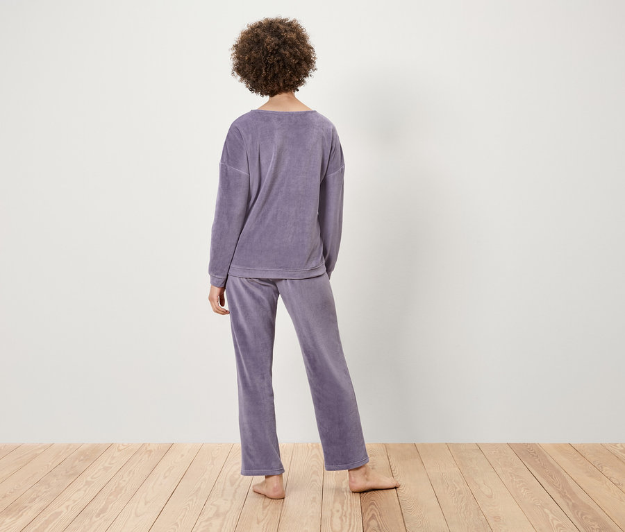 Kıvırcık saçlı bir kadın, açık renkli ahşap bir yüzey üzerinde mor kadife bir pijama giyerek arkası dönük duruyor.