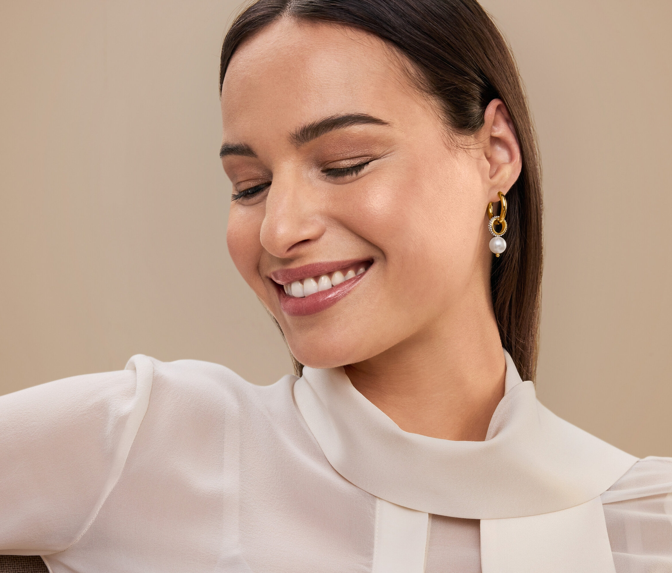 Kahverengi saçlı ve inci detaylı altın küpeli, beyaz bluzlu gülen kadın.