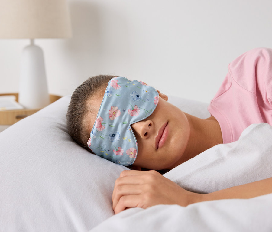 Çiçek desenli uyku maskesi takmış bir kadın yatakta uyuyor.