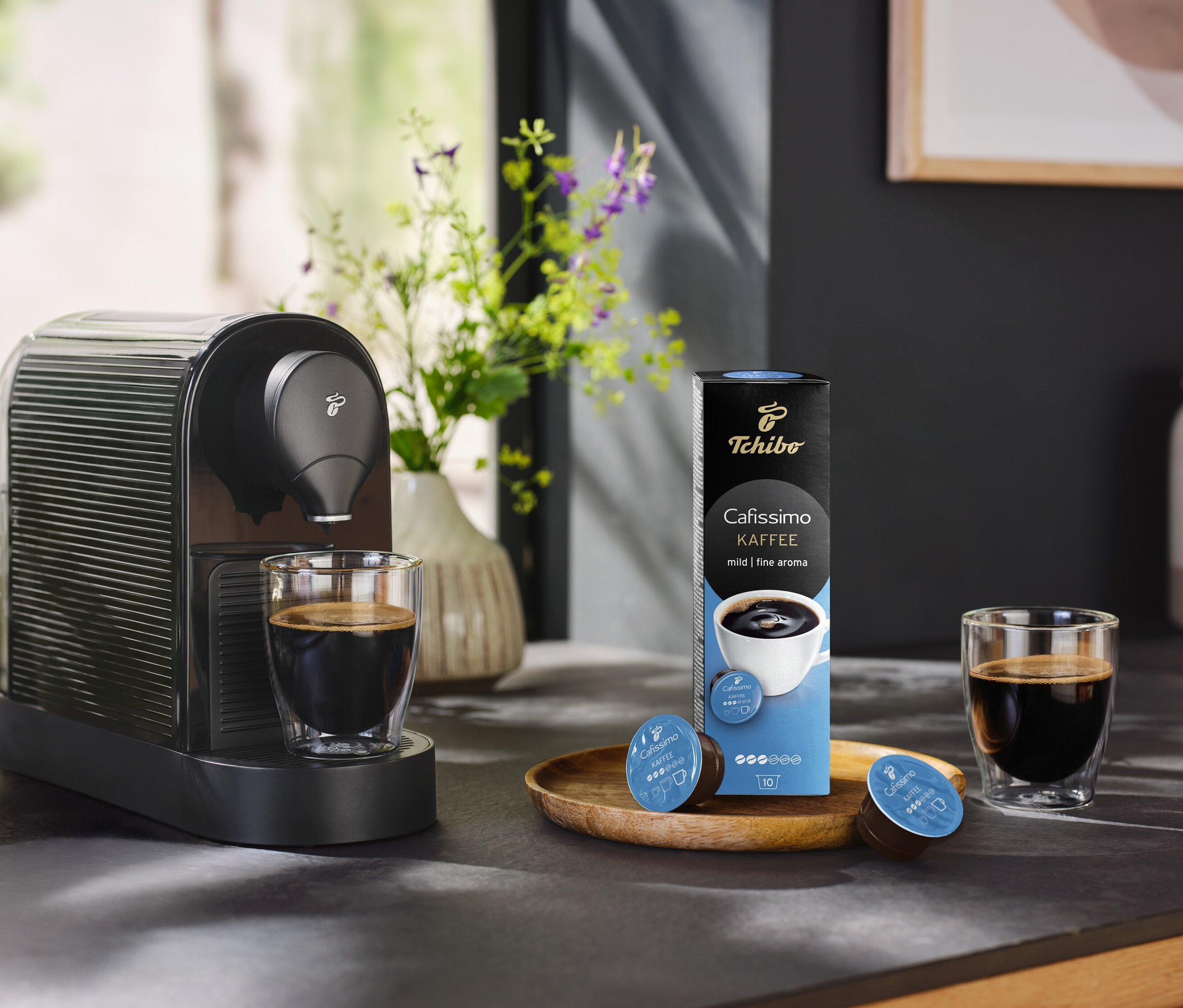 Kahve makinesi, bir bardak kahve ve bir paket 10 kapsül Coffee Fine Aroma Kapsül Filtre Kahve yanında duruyor.