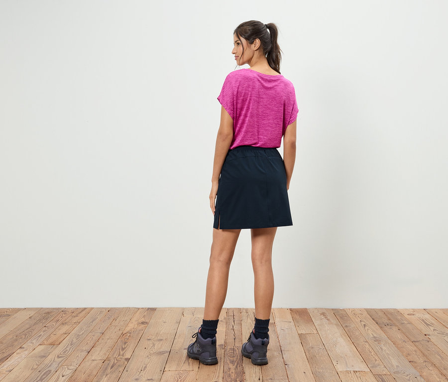 Atkuyruklu bir kadın arkası dönük duruyor, üzerinde pembe bir tişört, siyah bir etek ve gri ayakkabılar var.