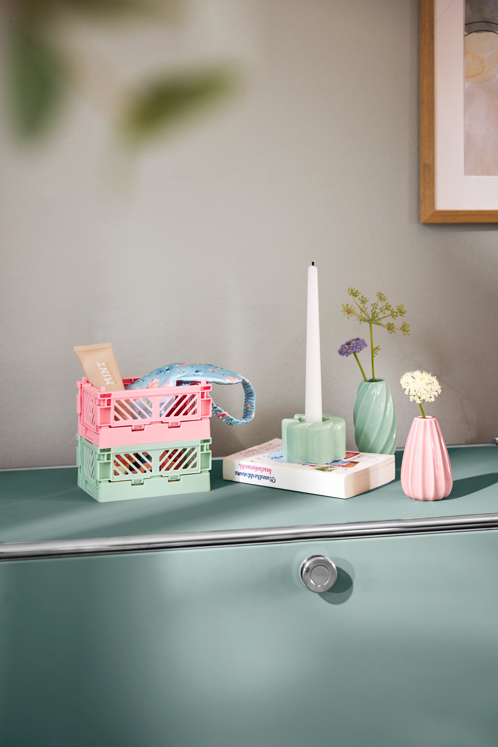 Adaçayı yeşili rengindeki, 1 adet açılır bölmesi olan metal yan dolap »CN3« üzerinde, pembe ve adaçayı yeşili renklerinde üst üste istiflenmiş, içinde bir el çantası ve bir tüp bulunan iki adet açık plastik kutu duruyor. Yanında bir kitap, mumlu bir mumluk ve çiçekli iki küçük vazo bulunuyor.