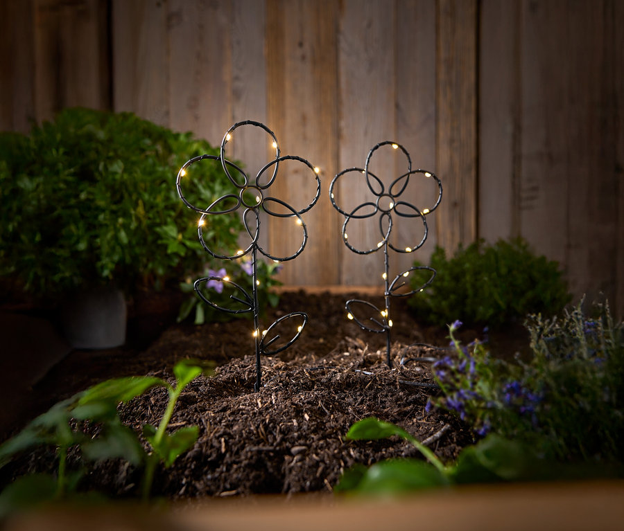 Ahşap bir duvarın önünde, toprakta LED ışıklı iki siyah, çiçek şeklinde bahçe kazığı duruyor.