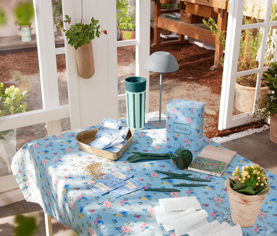 Çiçekli örtüyle kaplı masada gri LED akülü masa lambası, bahçe aletleri ve tohumlar duruyor.