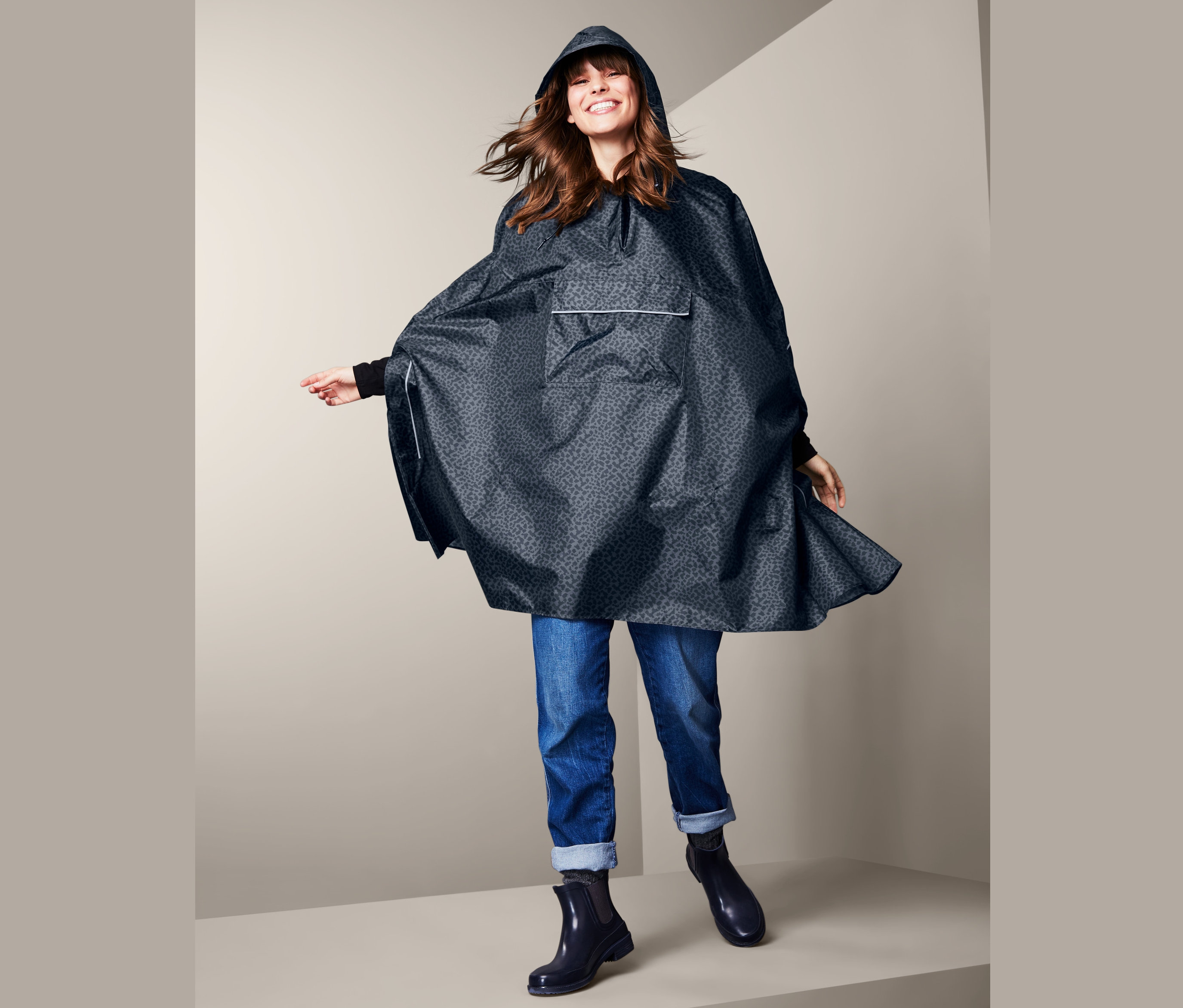 Gülümseyen bir kadın kapüşonlu mavi PANÇO YAĞMURLUK, kot pantolon ve botlarla poz veriyor.