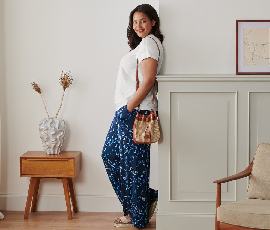 Bir kadın duvara yaslanmış, Palazzo Pantolon, beyaz tişört ve platform deri sandaletler giyiyor. Omzunda Büzgülü Çanta var.